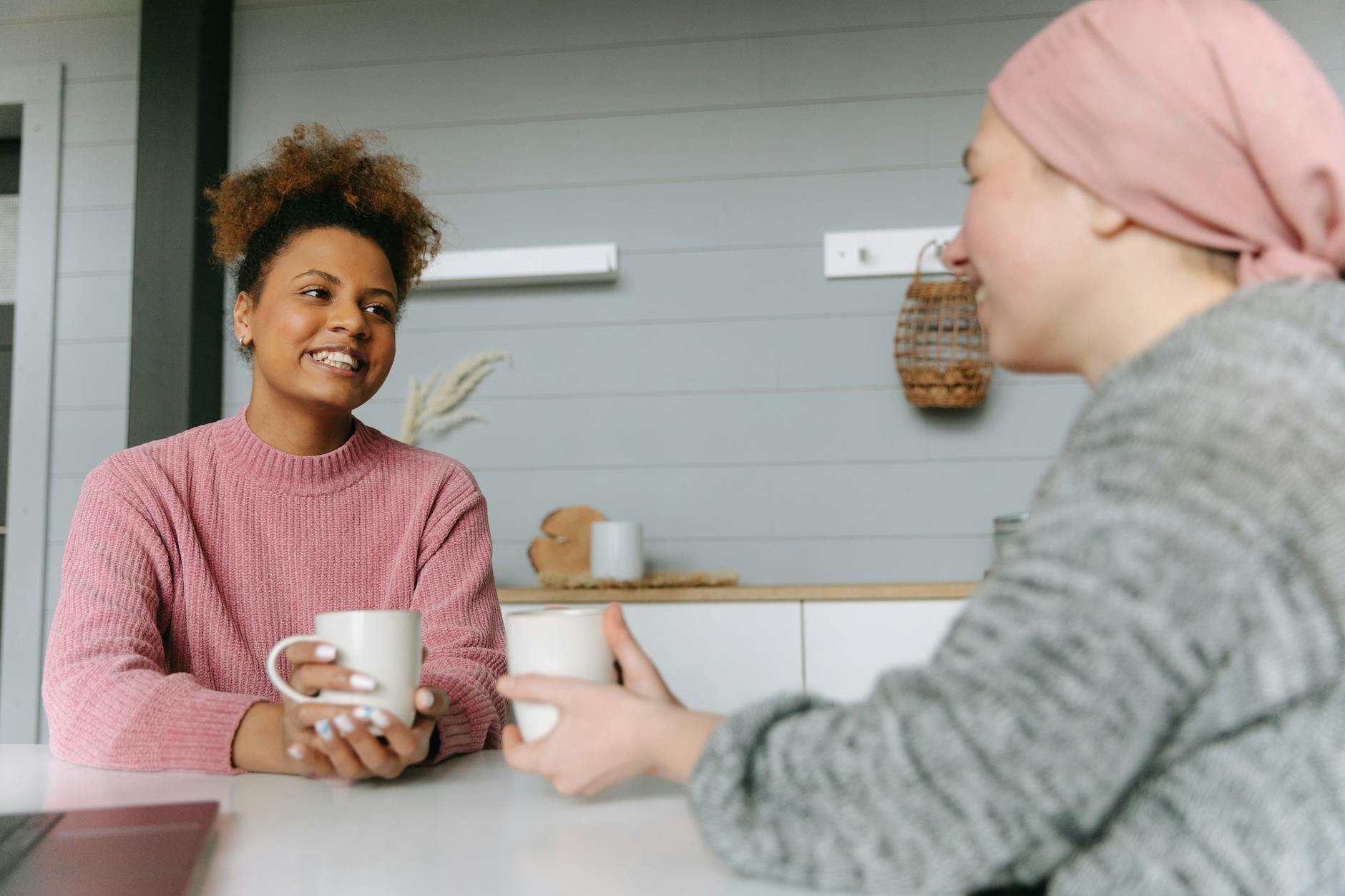 Two women sharing coffee and laughter at a kitchen table representing the everyday conversations ruled by the 3rd house