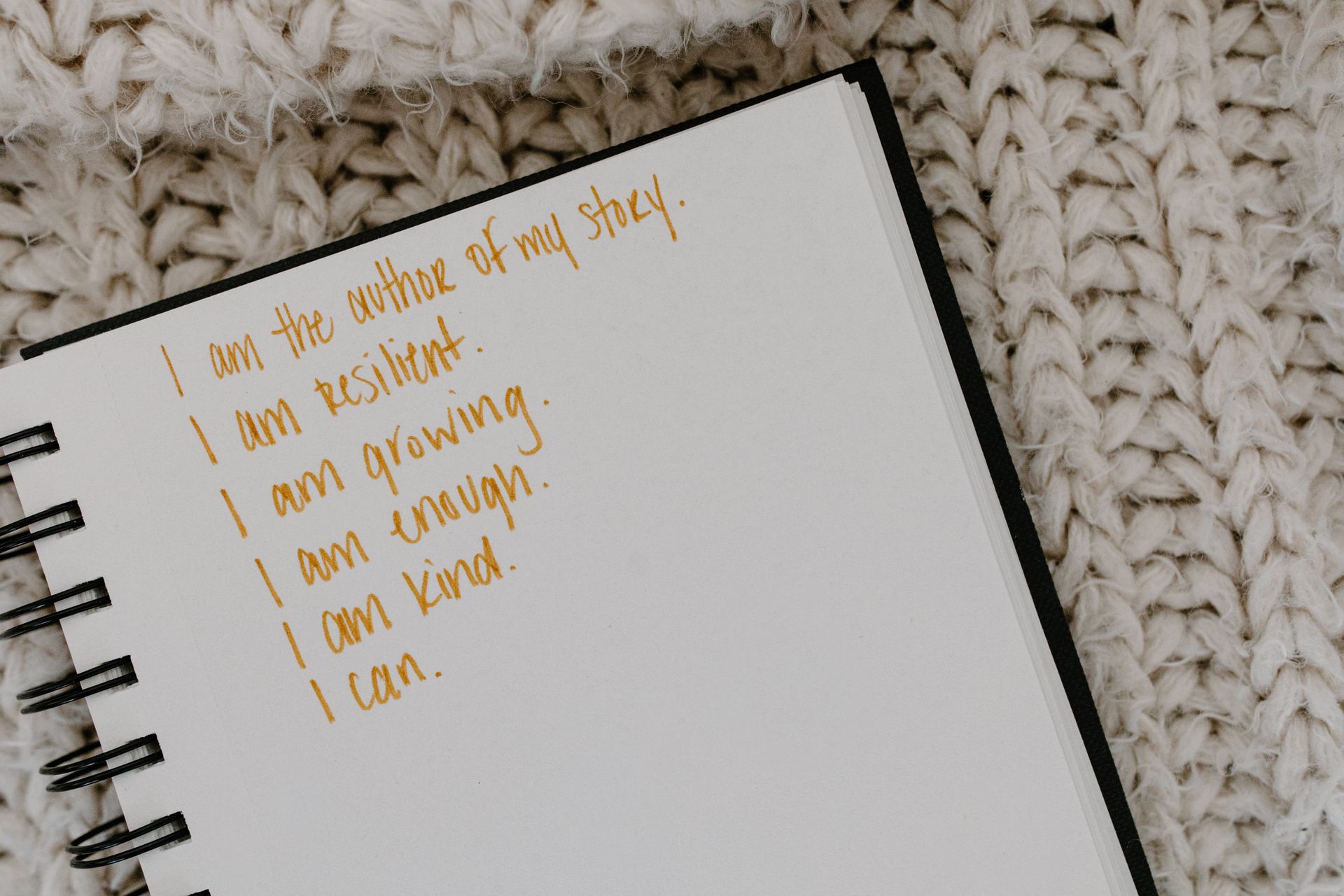 A spiral notebook on a knit blanket with handwritten affirmations in gold ink, the kind of positive statements written during the 55x5 method