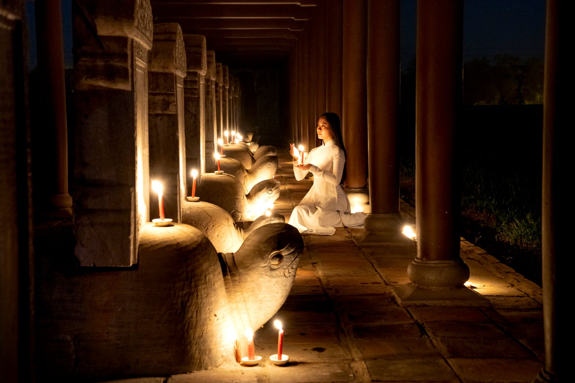 A woman in white kneeling with a candle in a candlelit temple corridor at night, representing the spiritual awakening and inner reflection connected to angel number 777