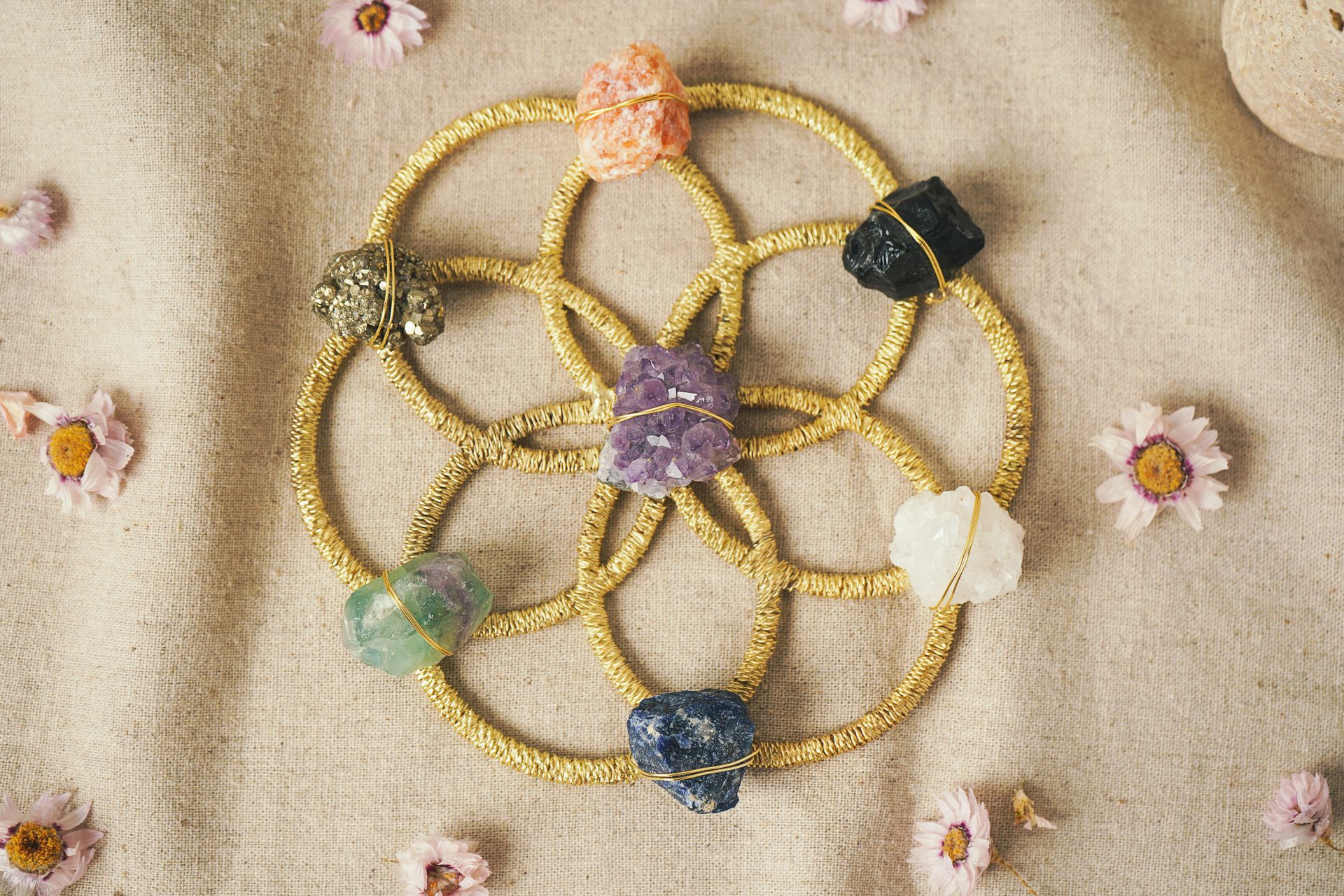 A crystal grid arranged on a golden Flower of Life pattern with amethyst at the center, surrounded by clear quartz, sodalite, fluorite, pyrite, and orange calcite on natural fabric