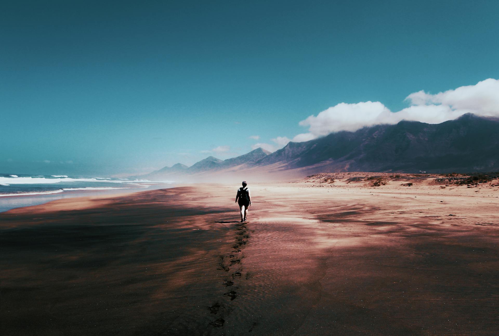 A lone person walking along a vast sandy beach with mountains in the background capturing the solitary journey of the Eight of Cups