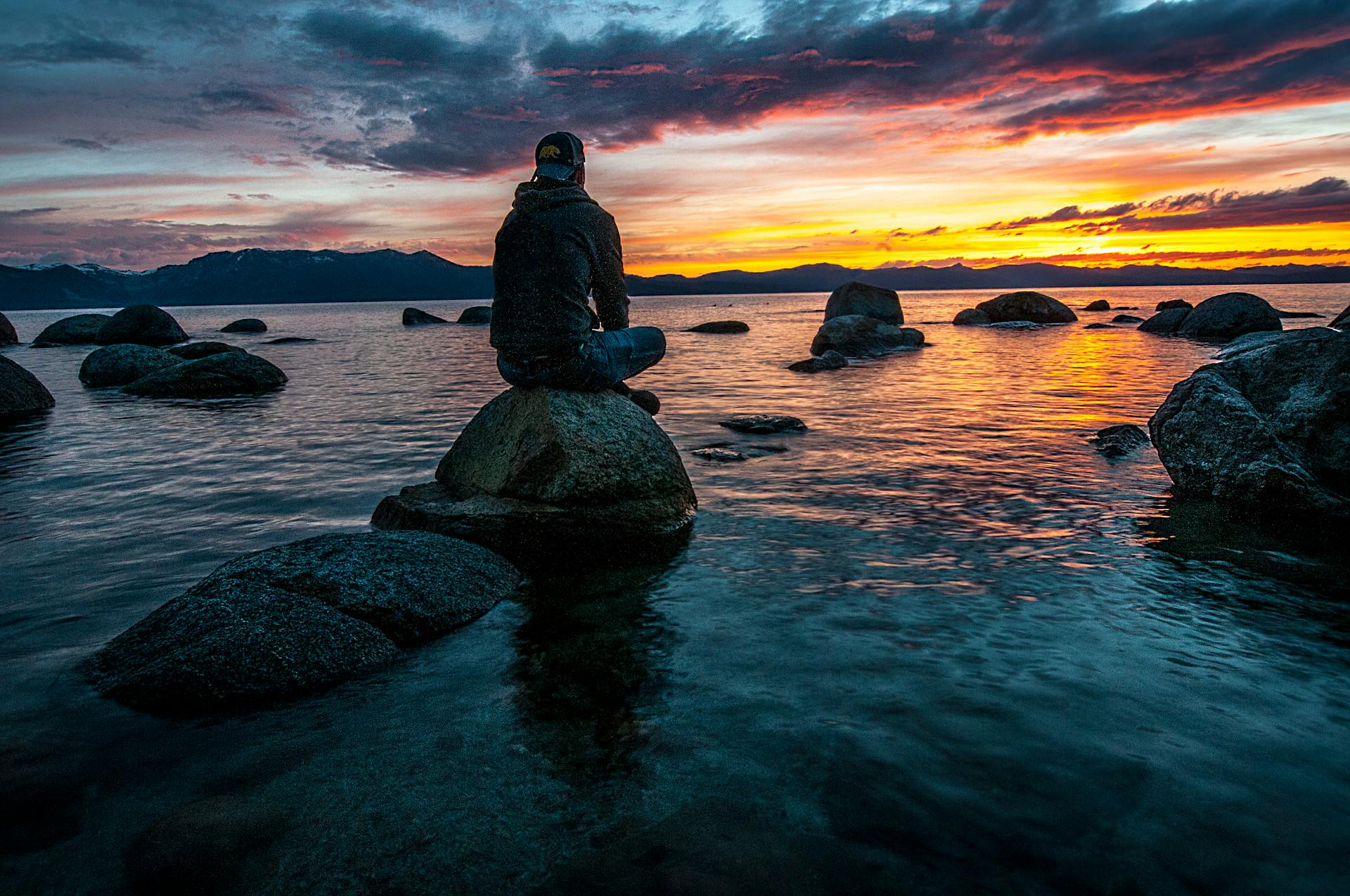 Silhouette of a man seated on rocks gazing at a serene lake sunset capturing the quiet emotional mastery and composed presence of the King of Cups