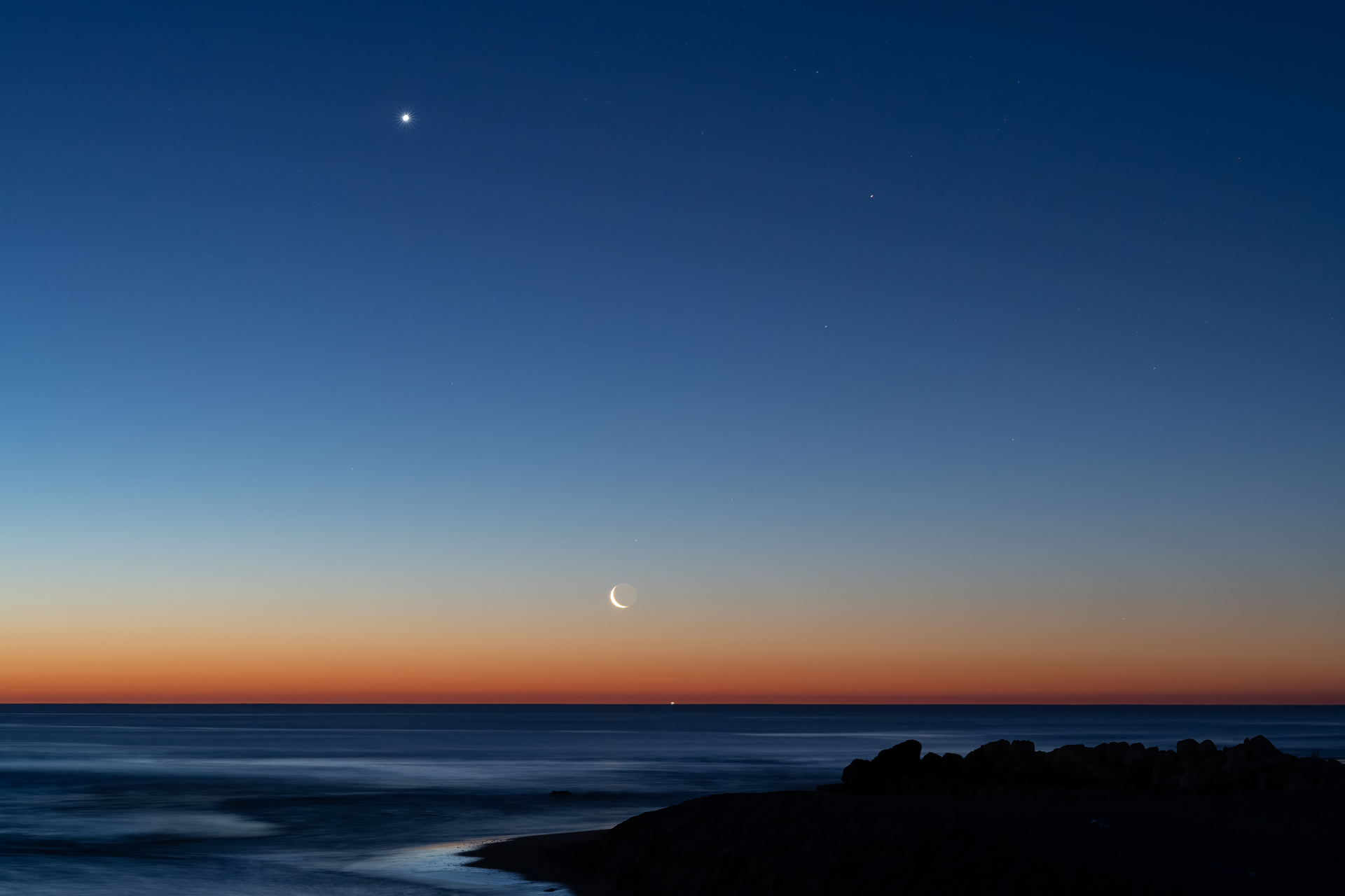 Crescent moon and planets aligning over the ocean at twilight, evoking two planets connected across the sky