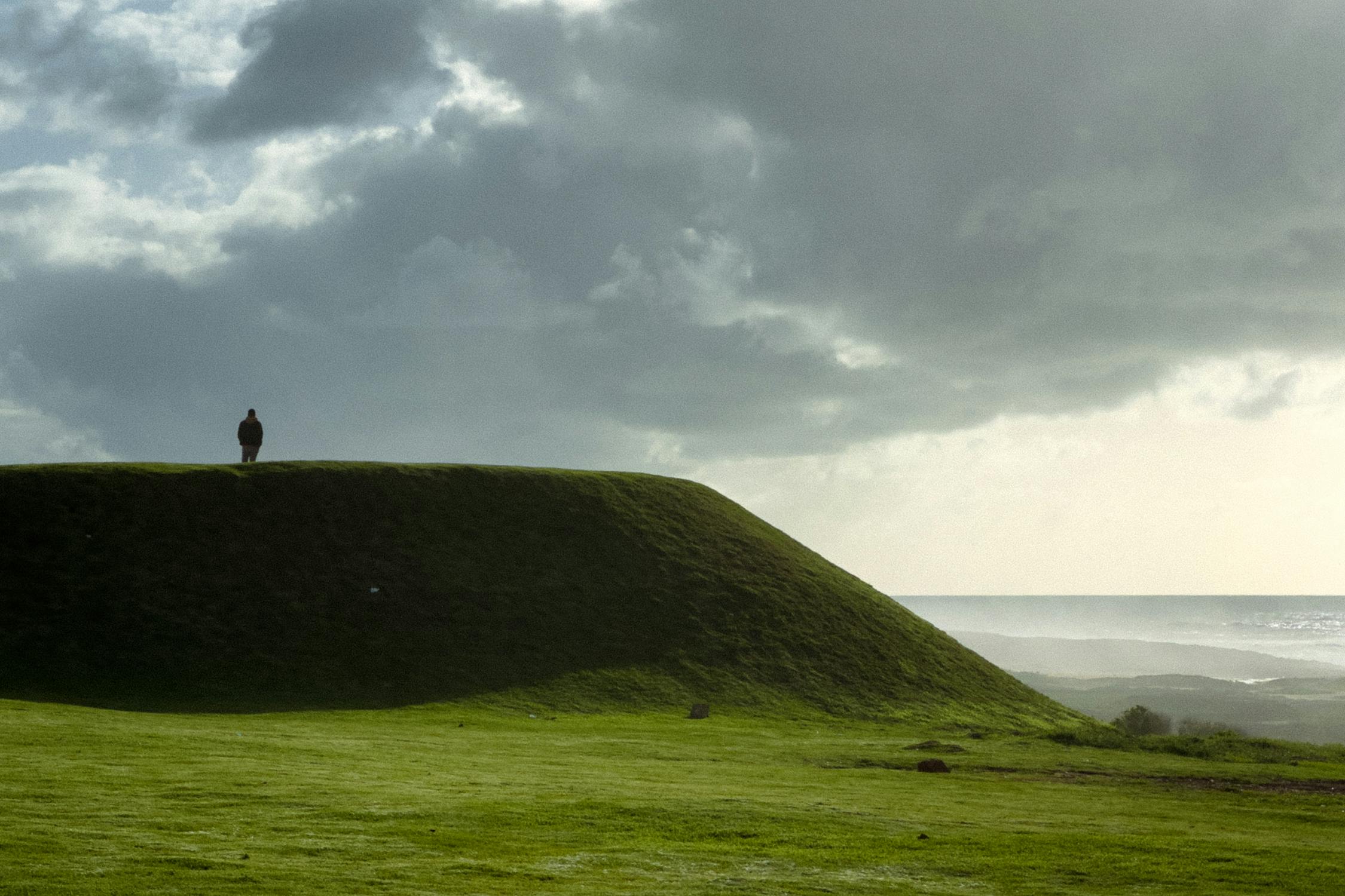 A solitary person on a grassy hill with an ocean view under dramatic clouds capturing the elevated watchful position of the Page of Swords surveying the intellectual landscape