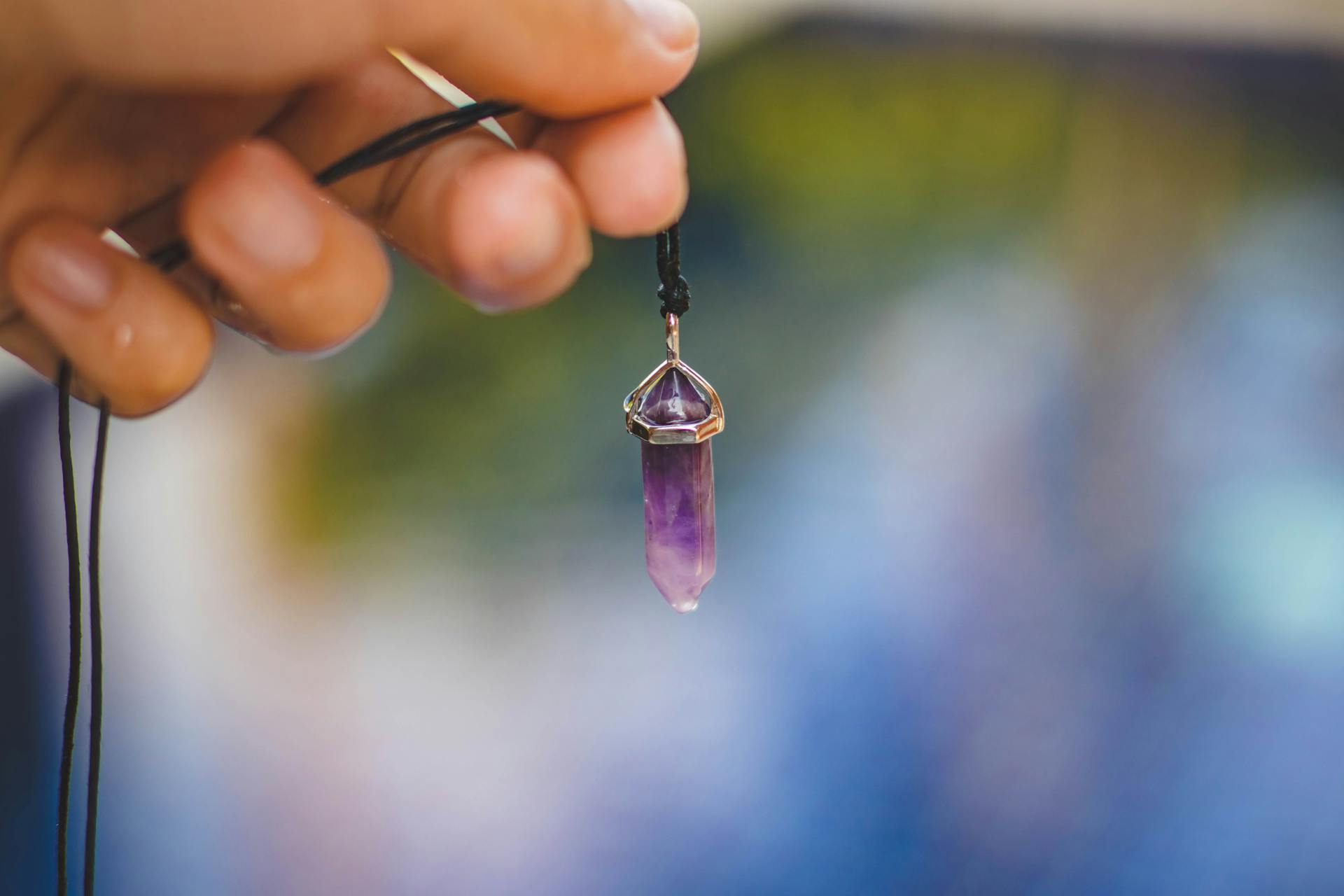 Close-up of a hand holding a purple amethyst crystal pendant on a chain against a natural outdoor background