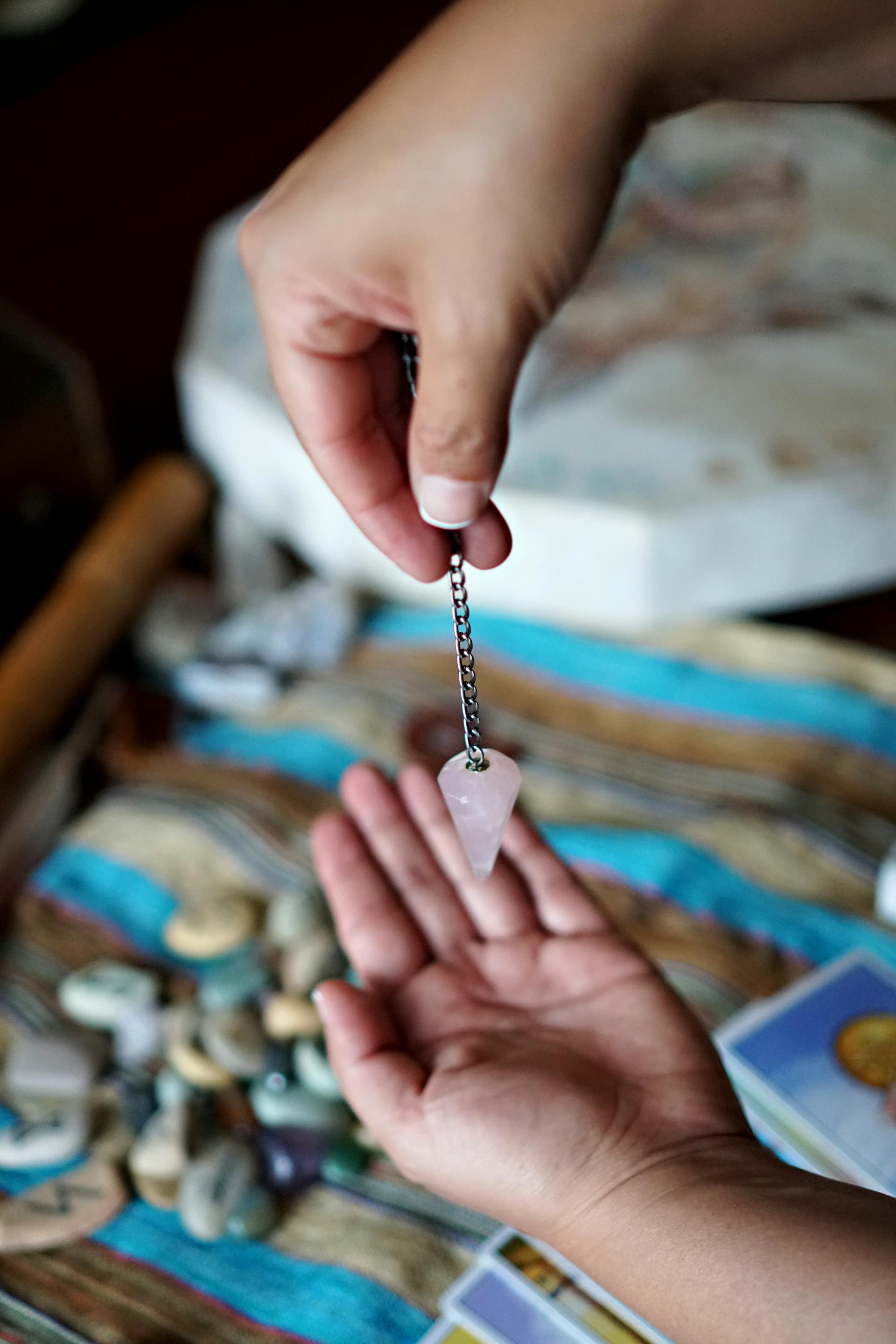 Two hands using a rose quartz pendulum over tarot cards spread on a table for divination