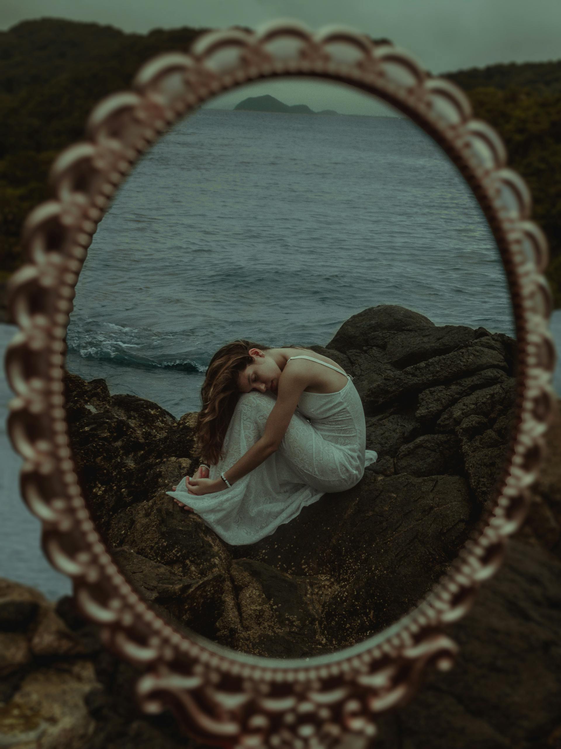 A woman in a white dress sitting reflectively on rocks by the ocean captured through a mirror showing the introspective emotional depth of the Queen of Cups
