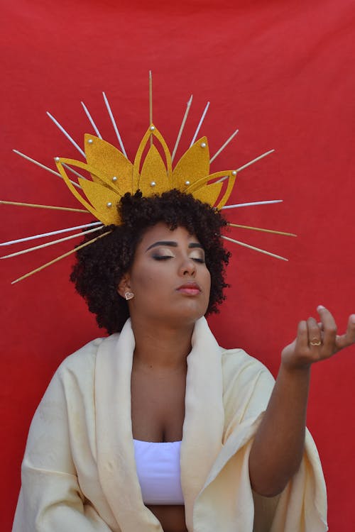 A woman wearing a radiant crown and white robe poses against a vivid red backdrop embodying the magnetic self-assurance of the Queen of Wands