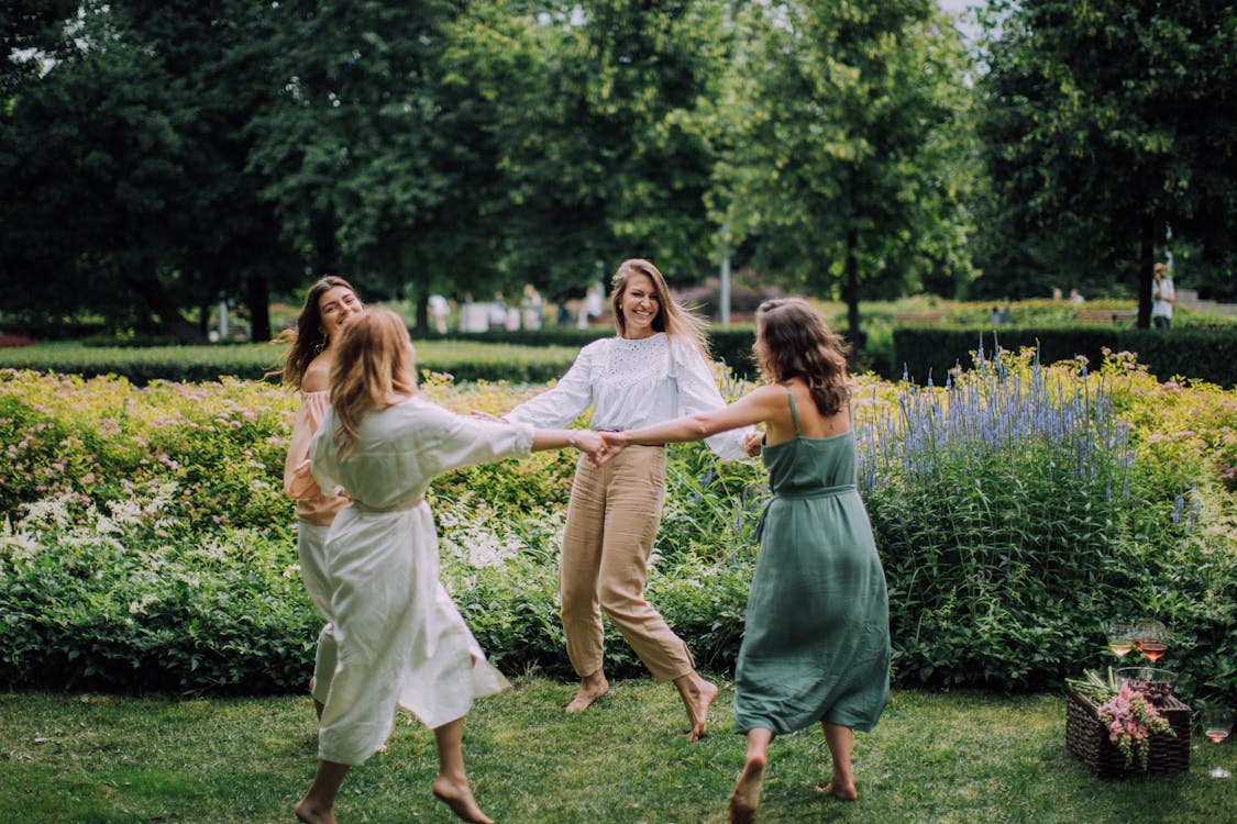 A group of women joyfully dancing together in a sun-drenched park radiating the spontaneous communal celebration that the Three of Cups represents