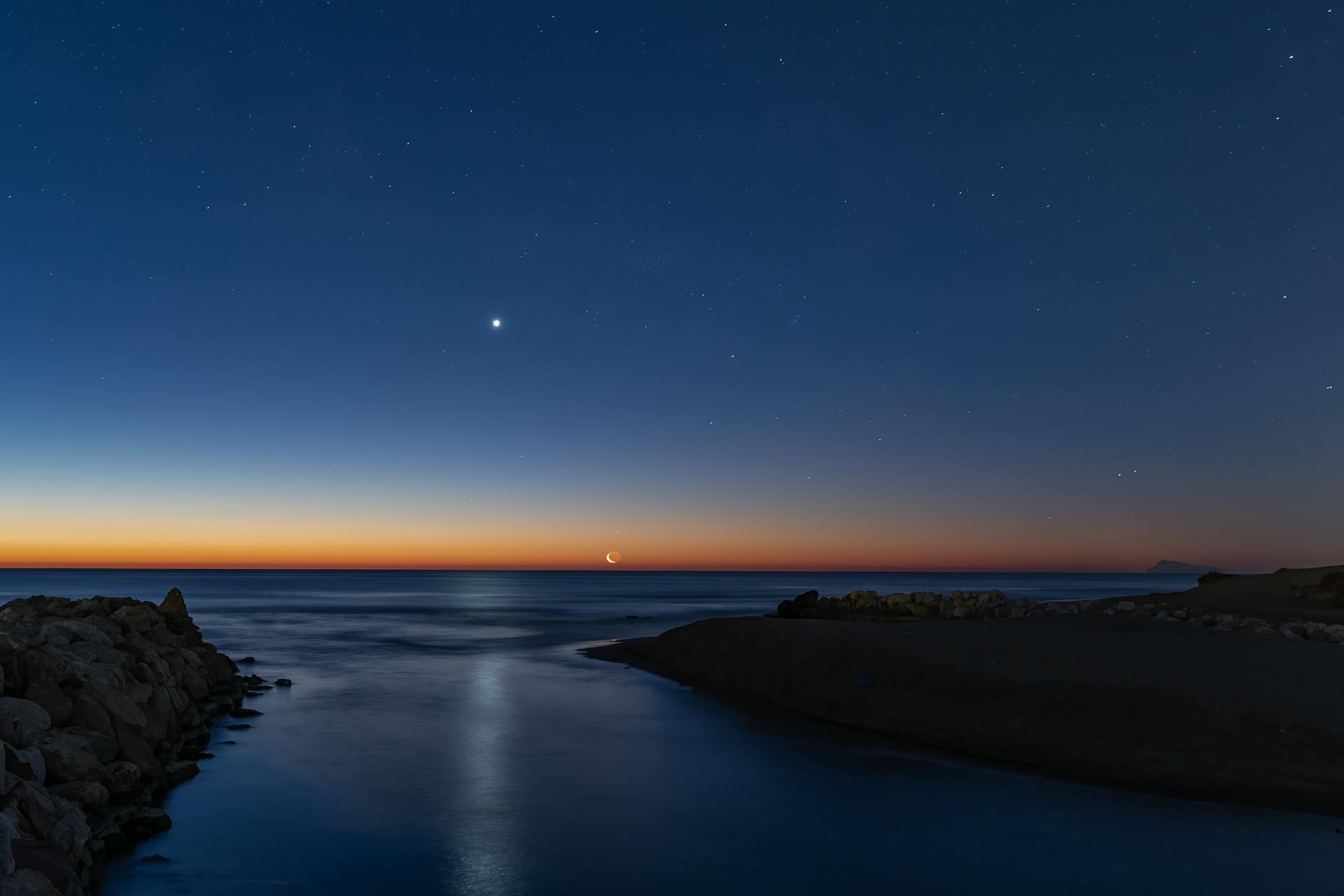 Crescent moon and bright star over a calm coastal seascape at twilight