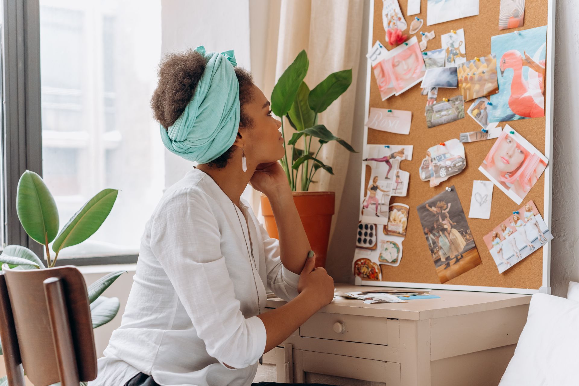 A woman in a white blouse sits at a desk looking at a corkboard vision board covered with pinned magazine images and photos, representing goals she wants to manifest